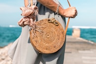 A woman wearing a checkered dress holds a woven round rattan bag with a patterned scarf tied to the handle. She is standing by the seaside, with the ocean in the background and a blue sky above.