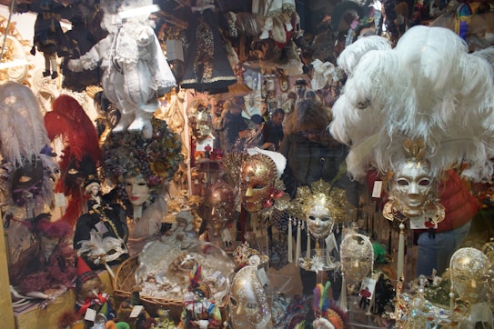 A vibrant and ornate display of Venetian masks and carnival costumes fills the scene. Elaborately designed masks with feathers, beads, and intricate patterns are prominently featured. Mannequin heads wear these masks, surrounded by plush dolls and other decorative items, creating a whimsical and festive atmosphere.