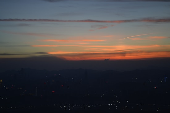 A cityscape silhouette is set against a stunning sunset, with vibrant hues of orange and red painting the sky above a darkened horizon. Wispy clouds are spread across the sky, adding texture to the scene.