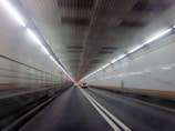 A well-lit tunnel with cars driving through it. The tunnel features white tiled walls and overhead lighting that creates a sense of motion. The perspective suggests speed and direction, with a clear focus on the lane markings and vanishing point.