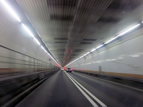 A well-lit tunnel with cars driving through it. The tunnel features white tiled walls and overhead lighting that creates a sense of motion. The perspective suggests speed and direction, with a clear focus on the lane markings and vanishing point.