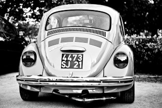 A vintage car with a classic design is parked outdoors, prominently displaying a license plate with the numbers 4473 SJ 21. The car is captured in grayscale, highlighting its distinctive round tail lights and the intricate detailing on the rear vent and bumper.