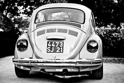 A vintage car with a classic design is parked outdoors, prominently displaying a license plate with the numbers 4473 SJ 21. The car is captured in grayscale, highlighting its distinctive round tail lights and the intricate detailing on the rear vent and bumper.