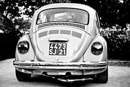 A vintage car with a classic design is parked outdoors, prominently displaying a license plate with the numbers 4473 SJ 21. The car is captured in grayscale, highlighting its distinctive round tail lights and the intricate detailing on the rear vent and bumper.