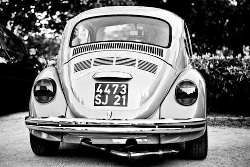 A vintage car with a classic design is parked outdoors, prominently displaying a license plate with the numbers 4473 SJ 21. The car is captured in grayscale, highlighting its distinctive round tail lights and the intricate detailing on the rear vent and bumper.