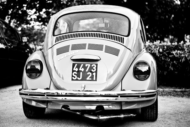 A vintage car with a classic design is parked outdoors, prominently displaying a license plate with the numbers 4473 SJ 21. The car is captured in grayscale, highlighting its distinctive round tail lights and the intricate detailing on the rear vent and bumper.