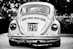 A vintage car with a classic design is parked outdoors, prominently displaying a license plate with the numbers 4473 SJ 21. The car is captured in grayscale, highlighting its distinctive round tail lights and the intricate detailing on the rear vent and bumper.