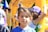 A young child wearing cat ears stands in the foreground, surrounded by colorful balloons and a vibrant, inflatable structure. The child has a neutral expression and is adorned with a beaded necklace. In the background, other people, including children, are engaging in activities, and the scene suggests a lively, festive outdoor event.