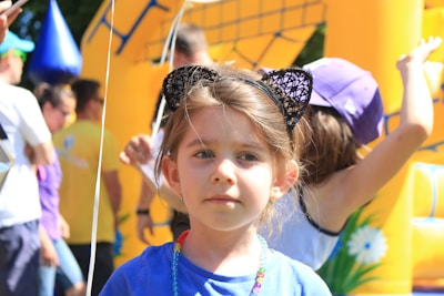 A young child wearing cat ears stands in the foreground, surrounded by colorful balloons and a vibrant, inflatable structure. The child has a neutral expression and is adorned with a beaded necklace. In the background, other people, including children, are engaging in activities, and the scene suggests a lively, festive outdoor event.