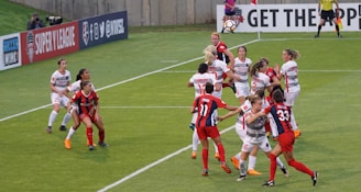 A group of female soccer players are actively competing for the ball during a match. Some players are jumping, indicating an aerial contest for possession. Team members are wearing different kits, with one team in red and blue uniforms and the other in white with red accents. The scene is dynamic and intense, emphasizing the competitive nature of the game. Advertisements and a referee are visible in the background, marking it as a professional sporting event.