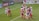 A group of female soccer players are actively competing for the ball during a match. Some players are jumping, indicating an aerial contest for possession. Team members are wearing different kits, with one team in red and blue uniforms and the other in white with red accents. The scene is dynamic and intense, emphasizing the competitive nature of the game. Advertisements and a referee are visible in the background, marking it as a professional sporting event.