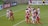 A group of female soccer players are actively competing for the ball during a match. Some players are jumping, indicating an aerial contest for possession. Team members are wearing different kits, with one team in red and blue uniforms and the other in white with red accents. The scene is dynamic and intense, emphasizing the competitive nature of the game. Advertisements and a referee are visible in the background, marking it as a professional sporting event.