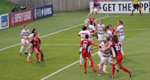 A group of female soccer players are actively competing for the ball during a match. Some players are jumping, indicating an aerial contest for possession. Team members are wearing different kits, with one team in red and blue uniforms and the other in white with red accents. The scene is dynamic and intense, emphasizing the competitive nature of the game. Advertisements and a referee are visible in the background, marking it as a professional sporting event.
