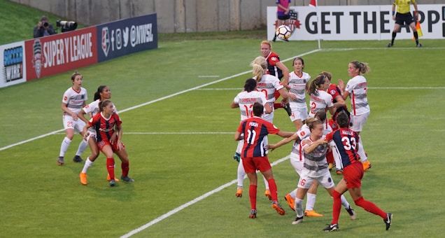 A group of female soccer players are actively competing for the ball during a match. Some players are jumping, indicating an aerial contest for possession. Team members are wearing different kits, with one team in red and blue uniforms and the other in white with red accents. The scene is dynamic and intense, emphasizing the competitive nature of the game. Advertisements and a referee are visible in the background, marking it as a professional sporting event.