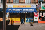 A storefront featuring services like money transfer, cheque cashing, and bureau de change. The awning prominently displays 'International Money Transfer' and various notices and advertisements are visible around the entrance. Adjacent to the entrance, fresh produce from a fruit vendor can be seen. The overall setting suggests a busy street, likely in an urban area.