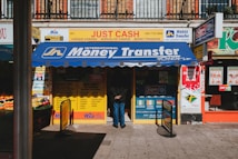 A storefront featuring services like money transfer, cheque cashing, and bureau de change. The awning prominently displays 'International Money Transfer' and various notices and advertisements are visible around the entrance. Adjacent to the entrance, fresh produce from a fruit vendor can be seen. The overall setting suggests a busy street, likely in an urban area.