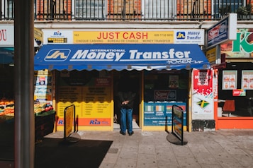 A storefront featuring services like money transfer, cheque cashing, and bureau de change. The awning prominently displays 'International Money Transfer' and various notices and advertisements are visible around the entrance. Adjacent to the entrance, fresh produce from a fruit vendor can be seen. The overall setting suggests a busy street, likely in an urban area.