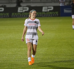 A female soccer player is walking on a grass field. She is wearing a white jersey with gray patterns and shorts, with orange Nike cleats. Her expression suggests a mix of exhaustion and disappointment.