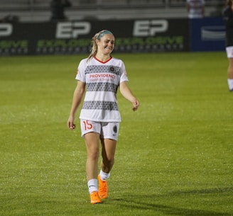 A female soccer player is walking on a grass field. She is wearing a white jersey with gray patterns and shorts, with orange Nike cleats. Her expression suggests a mix of exhaustion and disappointment.