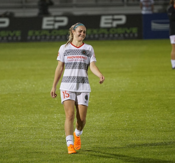 A female soccer player is walking on a grass field. She is wearing a white jersey with gray patterns and shorts, with orange Nike cleats. Her expression suggests a mix of exhaustion and disappointment.
