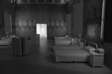 A room with multiple neatly arranged white beds, each paired with a small side table, creating a sense of symmetry. The walls feature geometric patterns, and there is a radiator placed near the doorway, which is illuminated from another room.