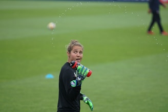 A person wearing a black long-sleeved shirt and green gloves sprays water from a red sports bottle. They are on a grass field, with another person blurred in the background. Water droplets are visible, creating an arc around the person.