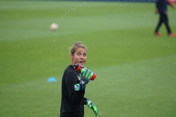 A person wearing a black long-sleeved shirt and green gloves sprays water from a red sports bottle. They are on a grass field, with another person blurred in the background. Water droplets are visible, creating an arc around the person.