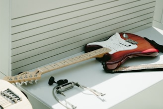 An electric guitar with a red and white body is resting on a white surface. Surrounding the guitar are various accessories including a guitar strap, a capo, and a set of earphones. The background features a series of horizontal slats that give a modern, sleek look.