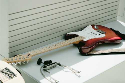 An electric guitar with a red and white body is resting on a white surface. Surrounding the guitar are various accessories including a guitar strap, a capo, and a set of earphones. The background features a series of horizontal slats that give a modern, sleek look.