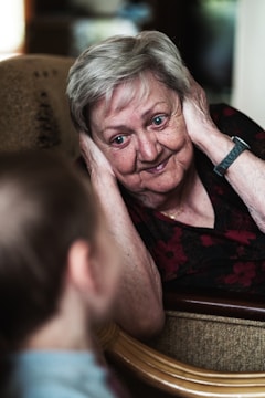 An elderly woman with short gray hair is leaning back on a chair, smiling warmly at a child in the foreground. She has her hands gently placed on the sides of her face, appearing engaged and delighted. The soft lighting and setting create an intimate and cozy atmosphere.