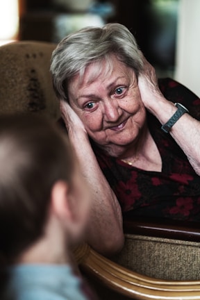 An elderly woman with short gray hair is leaning back on a chair, smiling warmly at a child in the foreground. She has her hands gently placed on the sides of her face, appearing engaged and delighted. The soft lighting and setting create an intimate and cozy atmosphere.
