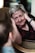 An elderly woman with short gray hair is leaning back on a chair, smiling warmly at a child in the foreground. She has her hands gently placed on the sides of her face, appearing engaged and delighted. The soft lighting and setting create an intimate and cozy atmosphere.