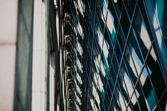 The image features the reflective surface of a modern building, displaying a grid of glass windows. The reflection creates abstract patterns with varying shades of blue and green, interspersed with lighter colors resembling the sky.
