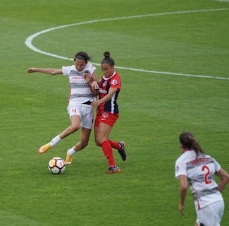 Two soccer players are engaged in a competitive match, vying for control of the ball on a well-maintained grass field. One player is wearing a white jersey with the number 4, while the other is in a red and blue jersey. Another player in a white jersey can be seen in the foreground.