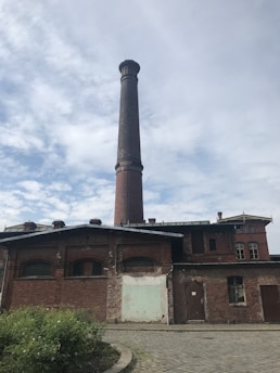 A tall, brick chimney rises from an old industrial building with a weathered appearance. The structure is made of red brick, featuring arched windows and a partially worn facade. Patches of greenery border the cobblestone path in the foreground. The sky is mostly cloudy with patches of blue.