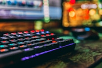 A close-up view of a colorful backlit mechanical keyboard with a blurred background. The keys are lit with vibrant red, blue, green, and purple lights, creating a visually striking effect. A computer mouse and a monitor with abstract, colorful bokeh can also be seen in the background.