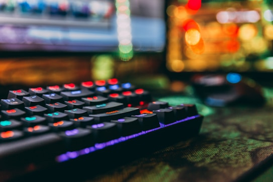 A close-up view of a colorful backlit mechanical keyboard with a blurred background. The keys are lit with vibrant red, blue, green, and purple lights, creating a visually striking effect. A computer mouse and a monitor with abstract, colorful bokeh can also be seen in the background.