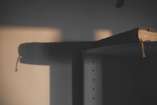 A dimly lit scene with a wooden shelf partially in shadows. A thick book with a textured cover and a bookmark hanging over the edge sits atop the shelf. Soft lighting casts rectangular shadows against a wall, creating an abstract pattern.