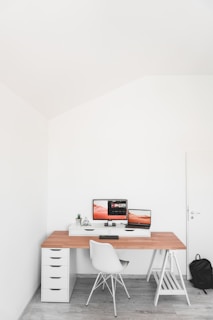 A minimalist workspace featuring a light wooden desk with a white chair. Two computer monitors display an orange-themed desktop background and editing software. The desk includes a small drawer unit on the left and a geometric decor piece along with a small potted plant. A backpack is placed on the floor to the right, and the room has a bright, clean aesthetic with white walls and light gray flooring.