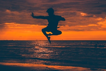 A silhouette of a person performing a high jump or martial arts kick in front of a vibrant sunset over the ocean. The sky is painted in shades of orange and red, with a few scattered clouds. The figure is captured mid-air against the backdrop of the setting sun, reflecting off the water's surface.