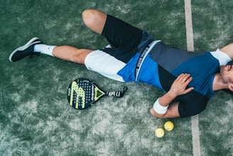 A person is lying on the ground, wearing athletic clothing that includes a blue and black top and white shorts. Nearby, there is a paddle racket and two yellow balls on a textured surface, which looks like a sports court. The person is wearing black sneakers and has a wristband on their arm.