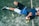 A person is lying on the ground, wearing athletic clothing that includes a blue and black top and white shorts. Nearby, there is a paddle racket and two yellow balls on a textured surface, which looks like a sports court. The person is wearing black sneakers and has a wristband on their arm.
