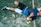 A person is lying on the ground, wearing athletic clothing that includes a blue and black top and white shorts. Nearby, there is a paddle racket and two yellow balls on a textured surface, which looks like a sports court. The person is wearing black sneakers and has a wristband on their arm.