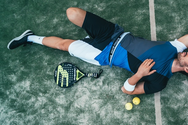 A person is lying on the ground, wearing athletic clothing that includes a blue and black top and white shorts. Nearby, there is a paddle racket and two yellow balls on a textured surface, which looks like a sports court. The person is wearing black sneakers and has a wristband on their arm.