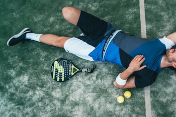 A person is lying on the ground, wearing athletic clothing that includes a blue and black top and white shorts. Nearby, there is a paddle racket and two yellow balls on a textured surface, which looks like a sports court. The person is wearing black sneakers and has a wristband on their arm.