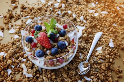 A glass bowl filled with granola, fresh strawberries, blueberries, and red currants is surrounded by scattered granola and coconut flakes on a wooden surface. A sprig of mint is placed on top of the fruit, adding a touch of greenery. A shiny spoon lies beside the bowl, ready for use.