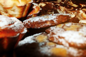 A close-up of various baked goods, including muffins and cookies, sprinkled with powdered sugar. The items appear freshly baked with a golden brown crust.