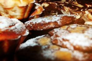 A close-up of various baked goods, including muffins and cookies, sprinkled with powdered sugar. The items appear freshly baked with a golden brown crust.