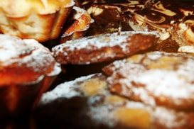 A close-up of various baked goods, including muffins and cookies, sprinkled with powdered sugar. The items appear freshly baked with a golden brown crust.