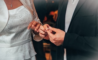 Two individuals participate in a wedding ceremony. The person on the left is dressed in a white gown with delicate lace and sequin details, while the person on the right is wearing a dark suit. They are exchanging rings, symbolizing a marriage commitment.